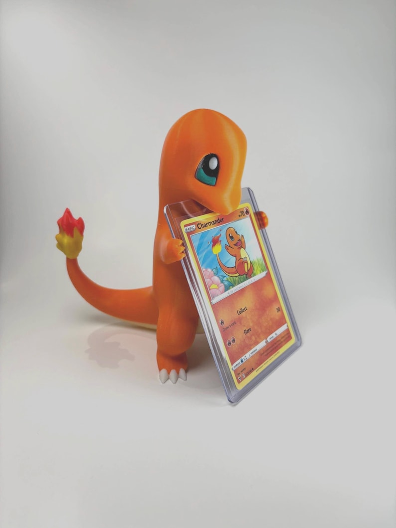May include: An orange Charmander figure holding a Charmander trading card in a protective case. The figure has a flame-shaped tail with red and yellow accents. The card features a colorful illustration of Charmander.