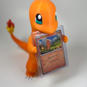 May include: A bright orange 3D-printed Charmander figure holding a Charmander Pok&eacute;mon trading card in a protective sleeve. The card shows an illustration of Charmander and the text "Blazing Destruction" and "Steady Firebreathing".