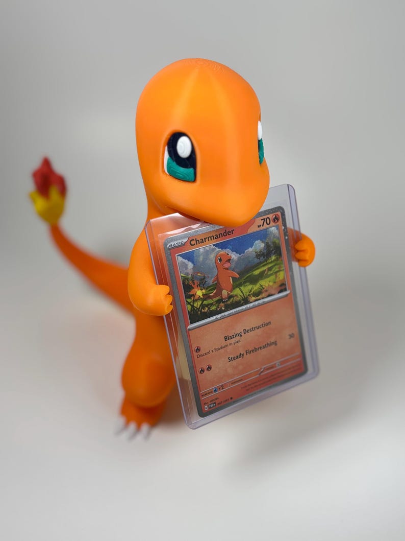 May include: A bright orange 3D-printed Charmander figure holding a Charmander Pok&eacute;mon trading card in a protective sleeve. The card shows an illustration of Charmander and the text "Blazing Destruction" and "Steady Firebreathing".