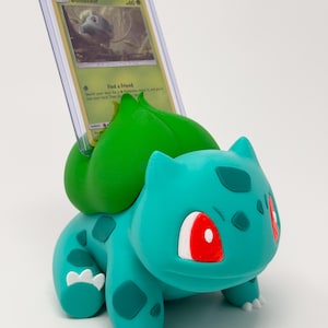 Op de afbeelding: Een turquoise Bulbasaur-figuur met een Pokémon-kaart. De figuur heeft rode ogen en groene accenten. De kaart zit in een beschermhoes en heeft een Bulbasaur-illustratie. Het is een verzamelobject.