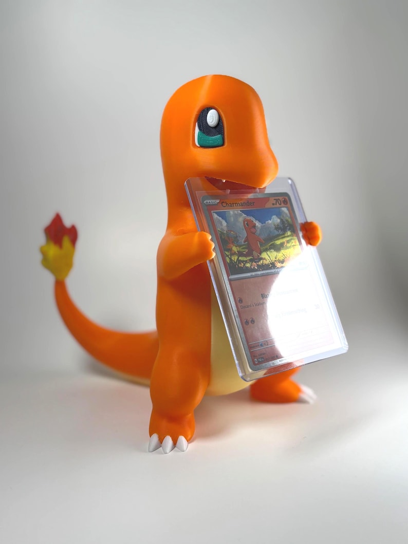 May include: An orange Charmander figurine holding a Pok&eacute;mon trading card in a protective case. The figurine has white claws, a yellow belly, and a tail with a red and yellow flame. The card features the name "Charmander".