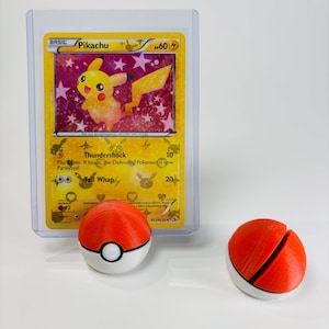 May include: A Pikachu Pokémon trading card in a protective case, with two red and white Poké Ball replicas. The card features a yellow Pikachu character with the text "Pikachu" and "60". The Poké Balls are red with a white band.