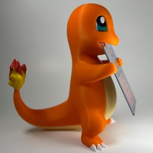 May include: An orange Charmander figure, holding a trading card. The figure has a yellow belly, white claws, and a flame-tipped tail. The trading card is in a protective sleeve. The background is a plain, neutral grey.