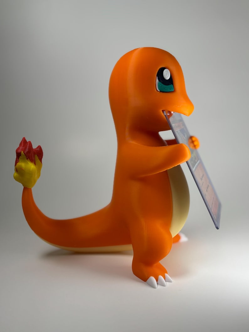 May include: An orange Charmander figure, holding a trading card. The figure has a yellow belly, white claws, and a flame-tipped tail. The trading card is in a protective sleeve. The background is a plain, neutral grey.