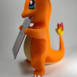 May include: A 3D-printed orange Charmander figure holding a trading card. The figure has white claws, a yellow belly, and a flame at the end of its tail. The eyes are black and blue. The figure is a collectible item.