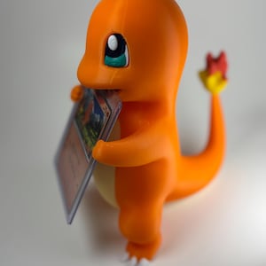 May include: A 3D-printed orange Charmander figure holding a trading card. The figure has white accents on its feet and a flame at the end of its tail. The card is in a protective sleeve.