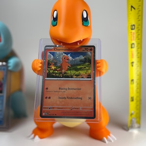 May include: An orange Charmander figure holding a protected Pok&eacute;mon trading card. The card features a Charmander illustration and the text "Blazing Destruction" and "Steady Firebreathing". A yellow measuring tape is in the background.