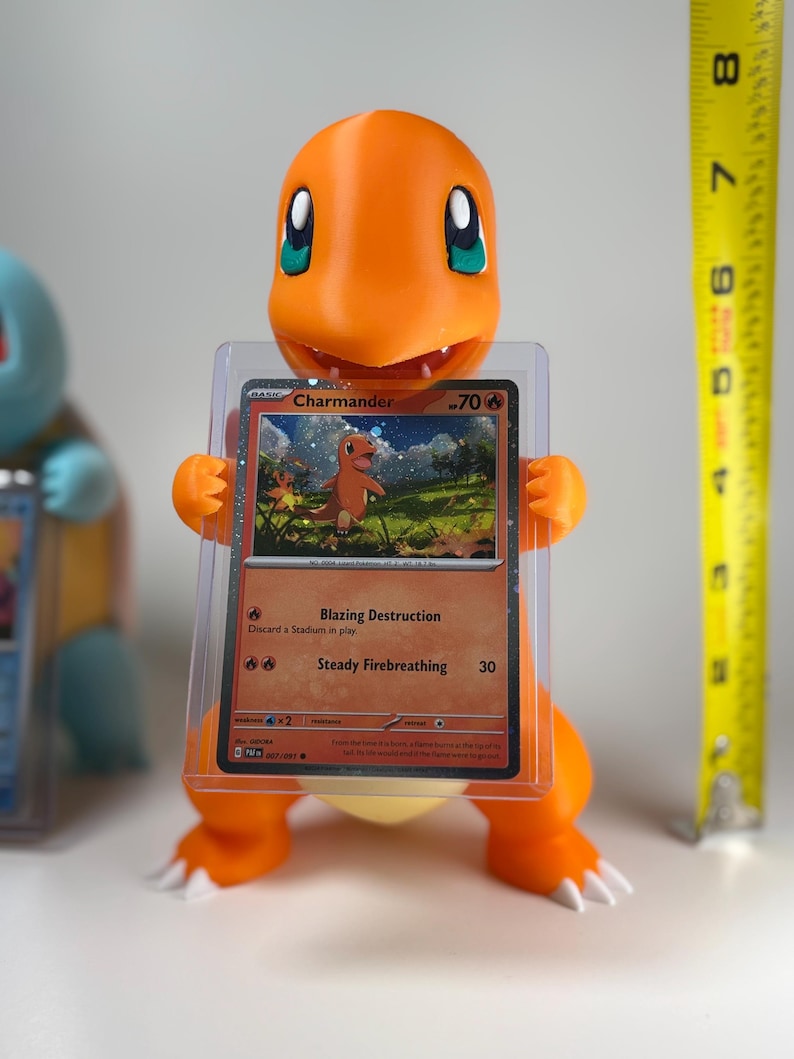 May include: An orange Charmander figure holding a protected Pok&eacute;mon trading card. The card features a Charmander illustration and the text "Blazing Destruction" and "Steady Firebreathing". A yellow measuring tape is in the background.