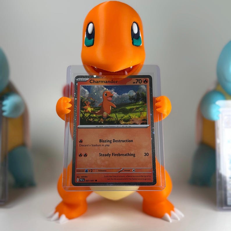 May include: A 3D-printed orange Charmander figure holding a Pok&eacute;mon trading card. The card displays a Charmander illustration with the text "Charmander" and "Blazing Destruction". The figure has white claws and a yellow belly.