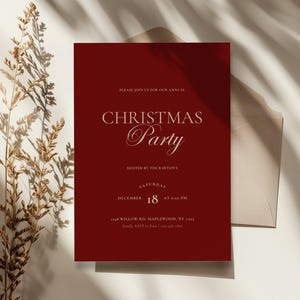 Puede incluir: Una invitación a una fiesta de Navidad roja con texto dorado. La invitación incluye las palabras "Christmas Party", la fecha, la hora y el lugar. Una planta seca y un sobre beige están en el fondo.