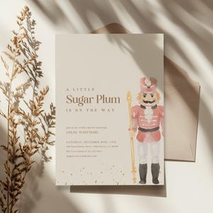 May include: A baby shower invitation with a watercolor illustration of a nutcracker soldier. The text reads "A Little Sugar Plum Is On The Way." The invitation is on a cream-colored background with gold accents.