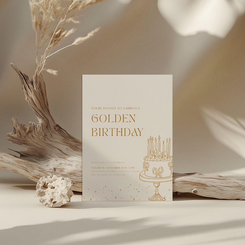 May include: A cream-colored invitation with gold text that reads "Golden Birthday." The invitation features a cake illustration with candles. Dried plants and a piece of driftwood are in the background.