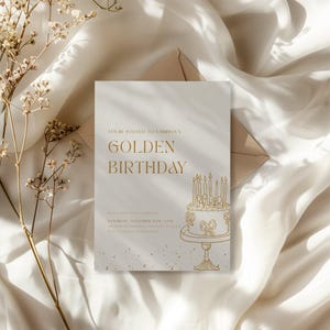 May include: A cream-colored invitation card with gold text that reads "Golden Birthday." The card features a gold illustration of a cake with candles. The invitation is on a white fabric background with dried flowers.