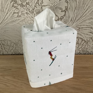 May include: Square white fabric tissue box cover with a skier design. The cover has a skier in red and yellow, skiing down a hill, with green dots and trees. The tissue box sits on a wooden surface.