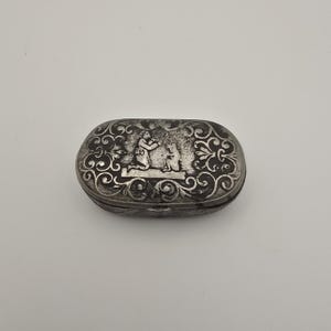 Puede incluir: Una tabaquera ovalada de metal con un acabado oscuro y deslustrado. La tapa presenta una escena en relieve de una figura arrodillada ante un animal, rodeada por un intrincado borde floral con volutas que cubre toda la superficie superior.