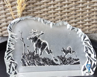 May include: A clear glass decorative piece featuring a moose and a dog in a naturalistic scene. The edges are jagged, and the design is etched into the glass. The piece has a rectangular shape with a textured background.