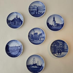 Può includere: Una collezione di sette piatti decorativi blu e bianchi. Ogni piatto presenta un'illustrazione scenica diversa, tra cui edifici, paesaggi e una nave. I piatti sono disposti su una parete bianca, creando un'esposizione.