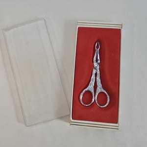 Pode incluir: Pinças de servir prateadas com detalhes ornamentados, apresentadas numa caixa forrada a vermelho com tampa transparente. A caixa tem uma borda listrada branca e dourada. As pinças têm uma pega em forma de tesoura.