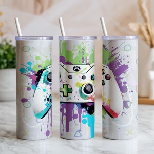 Puede incluir: Tres vasos de acero inoxidable con tapas y pajitas blancas. Cada vaso presenta un diseño colorido de un mando de Xbox con efectos de salpicaduras de pintura en verde, morado, azul y amarillo. Los vasos miden unos 20 cm de alto.