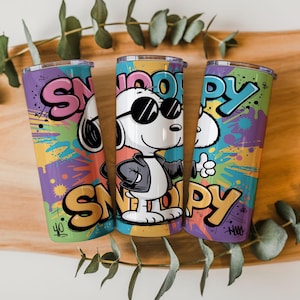 Könnte beinhalten: Drei Edelstahlbecher mit farbenfrohen Designs im Graffiti-Stil. Jeder Becher zeigt das Wort "Snoopy" in fetten Buchstaben und ein Cartoon-Bild von Snoopy mit Sonnenbrille. Der Hintergrund hat eine lebendige Mischung aus Farben, darunter Lila, Gelb und Blau.