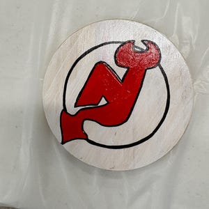 May include: A circular wooden coaster featuring a red and black logo. The logo is a stylized red "N" with a devil's tail and a black outline, within a black circle. The coaster has a natural wood grain finish.