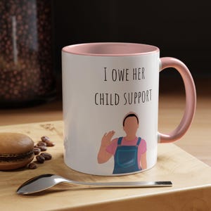 May include: White ceramic mug with a pink interior and handle. The mug features the text "I OWE HER CHILD SUPPORT" and an illustration of a person waving. A spoon and a macaron are in the foreground.