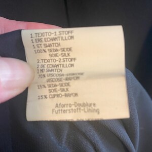 May include: A white fabric label with black text detailing the fabric composition of a garment. The label reads: "1.TEXITO-1.STOFF 1. ERE ECHANTILLON 1 ST SWATCH 100% SEDA-SEIDE SOIE-SILK 2.TEXITO-2.STOFF 2 DE ECHANTILLON 2 ND SWATCH 70% VISCOSA VISKOGE VISCOSE-RAPON 15% SEDA-SCIDE SOIE-SILK 15% CUPRO-RAYON Aforro-Doublure Futterstoff-Lining"