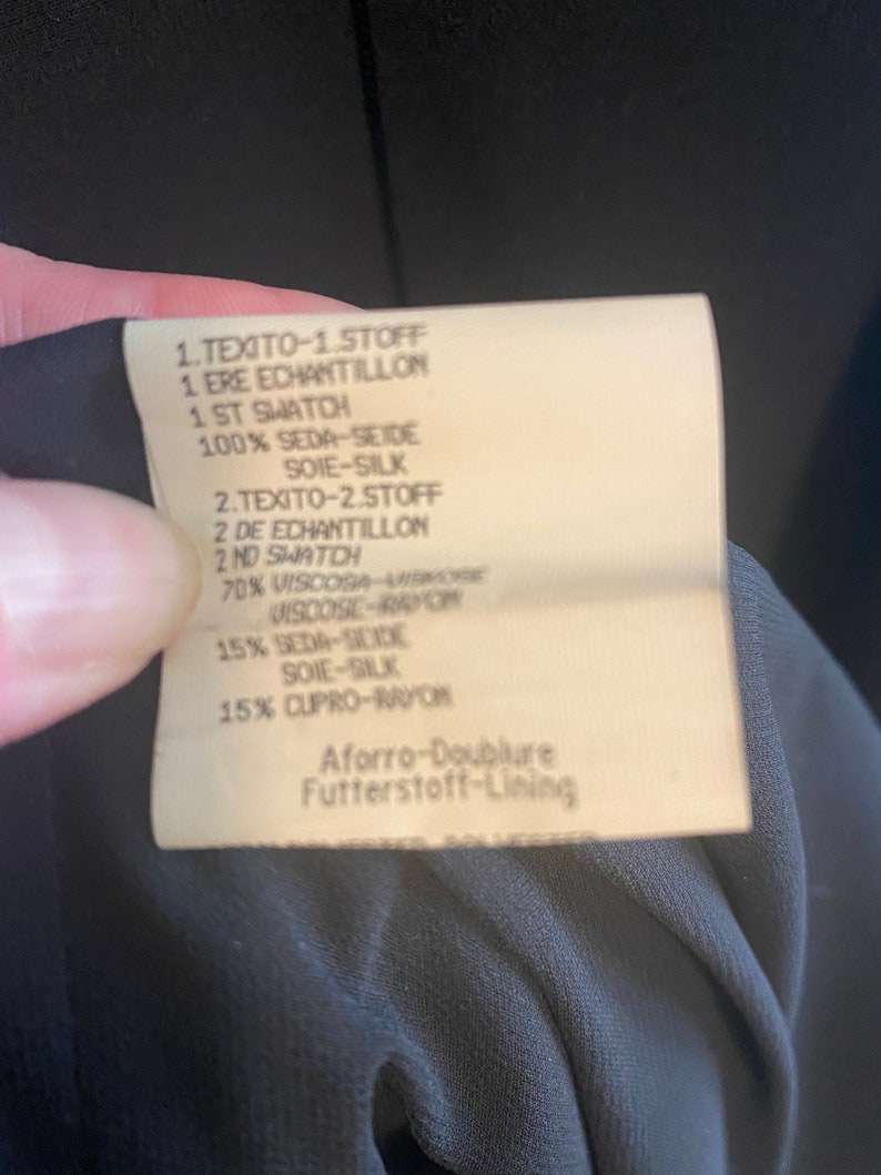 May include: A white fabric label with black text detailing the fabric composition of a garment. The label reads: "1.TEXITO-1.STOFF 1. ERE ECHANTILLON 1 ST SWATCH 100% SEDA-SEIDE SOIE-SILK 2.TEXITO-2.STOFF 2 DE ECHANTILLON 2 ND SWATCH 70% VISCOSA VISKOGE VISCOSE-RAPON 15% SEDA-SCIDE SOIE-SILK 15% CUPRO-RAYON Aforro-Doublure Futterstoff-Lining"