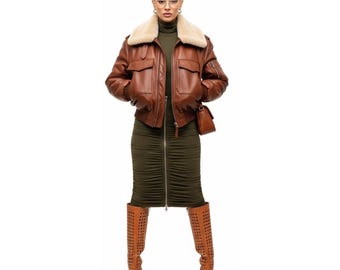 May include: A brown leather bomber jacket with a cream-colored shearling collar. The jacket has two front pockets and a small pocket on the left sleeve. The outfit includes a green dress and brown cutout boots.