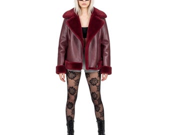 May include: A burgundy faux leather jacket with a shearling lining and trim. The jacket features a wide collar, a zipper closure, and a classic aviator style. The model is wearing black lace tights and black boots.