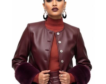 May include: A burgundy leather jacket with a round collar and three buttons. The jacket features faux fur cuffs in a matching burgundy color. The model is wearing a rust-colored turtleneck underneath the jacket.