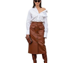 May include: A woman wearing a white button-down shirt and a brown, knee-length skirt with large pockets and buttons. She is also wearing sunglasses, a patterned clutch, and snakeskin-print boots.
