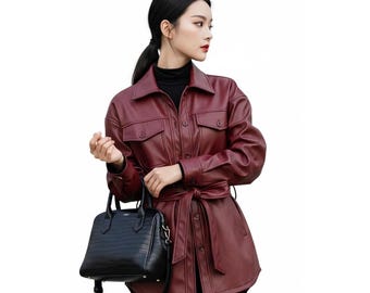 May include: A burgundy leather jacket with a collared neckline, two chest pockets, and a waist tie. The jacket is paired with a black handbag with a shoulder strap.