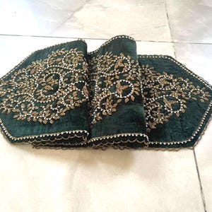 May include: A dark green velvet table runner with intricate gold and white beaded floral embroidery. The runner has a scalloped edge and decorative tassels on each end. The design is symmetrical and elegant.