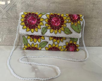 Può includere: Una pochette bianca con perline con una lunga tracolla bianca. La pochette è decorata con girasoli di perline gialle, rosse e rosa e foglie verdi. La borsa ha una forma rettangolare con chiusura a patta.