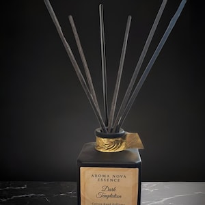 Puede incluir: Difusor de caña de algodón Aroma Nova Essence Dark Temptation. La botella cuadrada de vidrio negro tiene un acento dorado y contiene cañas negras. La etiqueta dice "Dark Temptation" y "4 oz". El difusor se encuentra sobre una superficie de mármol negro.