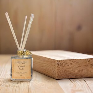 Puede incluir: Difusor de caña de algodón Aroma Nova Essence Earth & Ember con cañas. La botella de vidrio transparente tiene una cinta y una etiqueta doradas. Un bloque de madera está al lado del difusor. El fondo es una superficie de madera marrón claro.