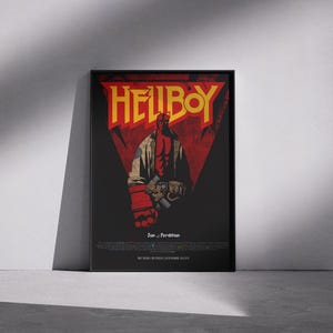 May include: A framed poster featuring the word "HELLBOY" in yellow, set against a red triangle on a black background. The poster depicts a red-skinned character with horns, holding a gun. The poster is leaning against a white wall.