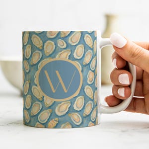 May include: White ceramic mug with a blue and gold oyster shell pattern. A light blue circle with a gold border and the letter "W" is centered on the mug. The mug is held by a person.