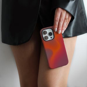 May include: A smartphone case with a gradient design in shades of orange, red, and purple. The case has a black border around the camera lenses. The phone case is held against a black leather jacket.