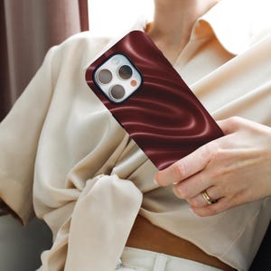 May include: A burgundy phone case with a flowing, satin-like design. The case has a black border around the camera and flash. The phone case is held in a person's hand.