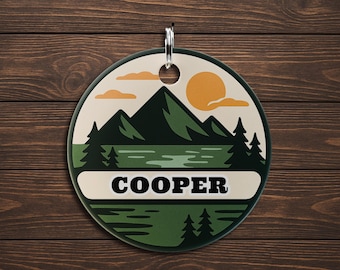May include: A round, green and cream-colored tag with a mountain and lake scene, a yellow sun, and the name "COOPER" in bold black letters. The tag has a silver ring for hanging and is set against a wood background.