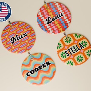 May include: Four colorful, round pet tags with retro designs and names: "Max", "Luna", "Stella", and "Cooper". Each tag has a different pattern in shades of orange, blue, yellow, and green. A metal ring is attached for easy attachment. The tags are made in the USA.