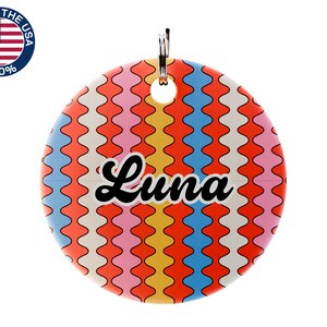 May include: A round pet tag with a colorful wavy pattern in orange, yellow, pink, and blue. The name "Luna" is written in black script across the center. A metal ring is attached at the top for easy attachment to a collar.
