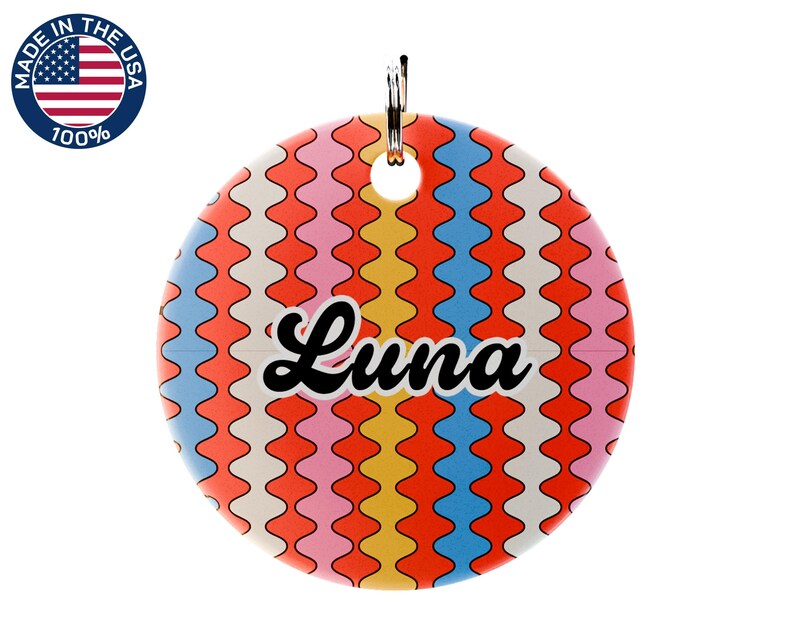 May include: A round pet tag with a colorful wavy pattern in orange, yellow, pink, and blue. The name "Luna" is written in black script across the center. A metal ring is attached at the top for easy attachment to a collar.