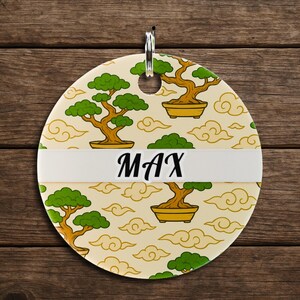 May include: A round, cream-colored tag with a bonsai tree and cloud pattern. The word "MAX" is printed in black on a white band across the center. A silver ring is at the top for hanging.