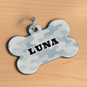 May include: A bone-shaped pet tag with a camouflage pattern in shades of light blue and white. The name "LUNA" is printed in bold black letters across the center. A silver-colored ring is attached at the top for easy attachment to a pet's collar.