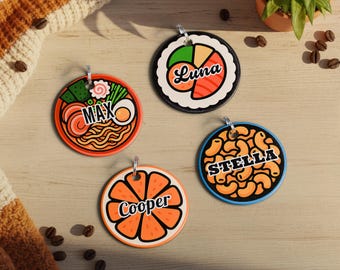 May include: Four round pet tags with food-themed designs. One tag features ramen with the name "MAX", another sushi with "Luna", a third with orange slices and "Cooper", and the last with macaroni and cheese and "Stella". Each tag has a silver loop.