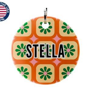 May include: A round, orange pet tag with a retro floral pattern in green and cream. The name "STELLA" is displayed in bold black letters. A metal ring is attached at the top. The tag is marked "MADE IN THE USA 100%."