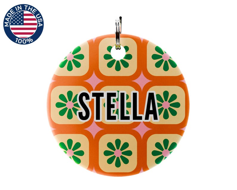 May include: A round, orange pet tag with a retro floral pattern in green and cream. The name "STELLA" is displayed in bold black letters. A metal ring is attached at the top. The tag is marked "MADE IN THE USA 100%."